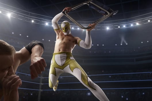 An image of two professional wrestlers in a match. One lunges over the other while the other seemingly cowers, protecting his head with his arm from a coming blow.
