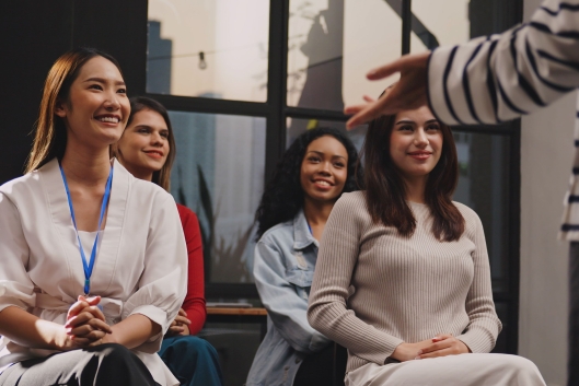 A group of four young women smile and listen attentively in the audience for a lecturer or seminar presenter.
