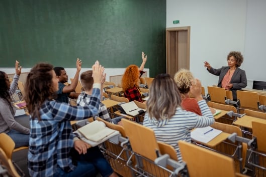 A diverse group of college students raise their hands as the instructor appears to call on one of them.