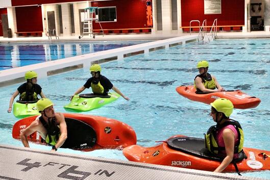 Five students sit in kayaks in an indoor swimming facility
