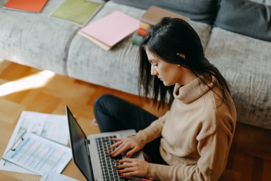 A young woman types on a laptop with papers strewn around her. 