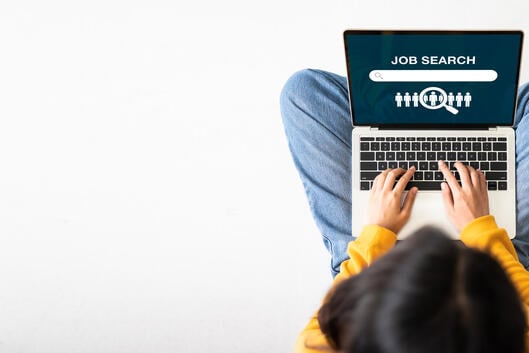 A young adult woman dressed casually in jeans and a sweater sits cross-legged on the floor, with a laptop open to a page that says "Job Search." The photo is taken from overhead.
