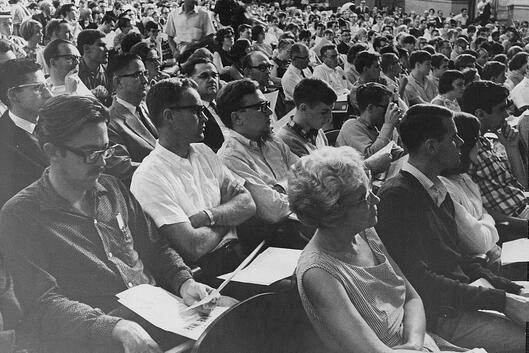 In this historical black-and-white photo from a 1965 anti-war teach-in, a dense crowd of people in an auditorium sit in an audience, seemingly listening to a speaker with intense interest and focus.