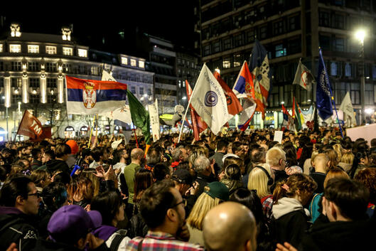 An image of a dense crowd of student protesters, some waving flags, at nighttime against the Belgrade cityscape.