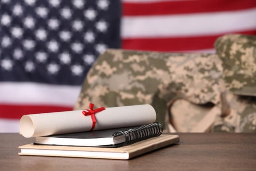 Two notebooks and a rolled-up diploma sit on a table in front of an empty chair draped with an Army jacket. The background of the photo is an American flag.
