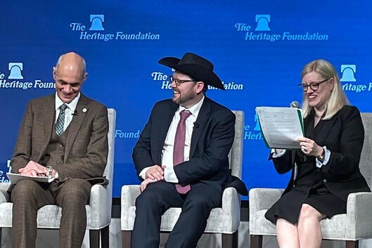 A photo of three panelists speaking at the Heritage Foundation. The man in the center is wearing a cowboy hat.
