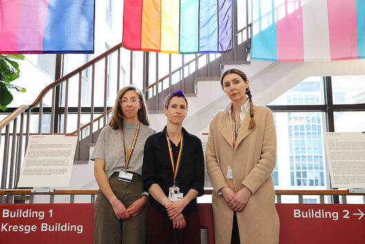 Instructor Ariel Beccia and associate professors Julia Marcus and Brittany Charlton pose in front of a series of LGBTQ flags.