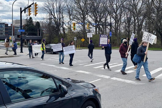 A photograph of demonstrators, holding signs, walking across a crosswalk.
