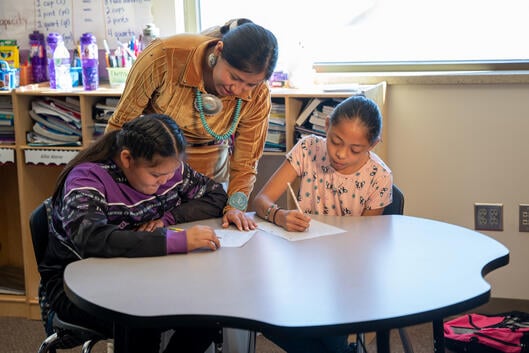 A teacher leans over a desk where two students are writing. 