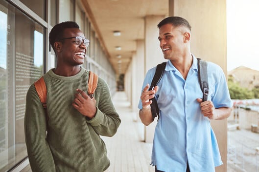 Two students wearing backpacks laugh while walking down a hallway at a college.
