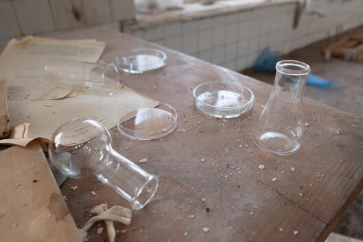 A photo of a dusty wooden table in an unused science laboratory; glass flasks, one knocked on its side, Petri dishes, and documents are strewn about the table. 