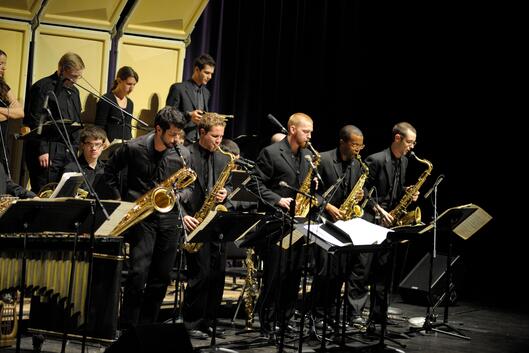 Five saxophone players stand to perform during a band concert. All students are clothed in black formal wear. 