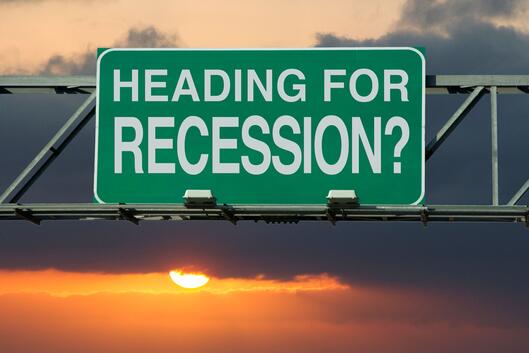 A green and white road sign, as if above a highway, says "Heading for Recession?" against the backdrop of a sunset in a cloudy sky.
