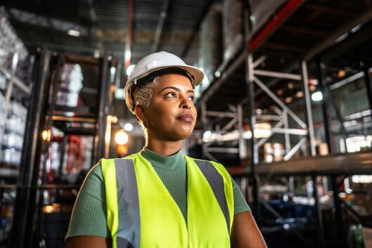 A Black woman in a hard hat and neon vest stands in a warehouse.