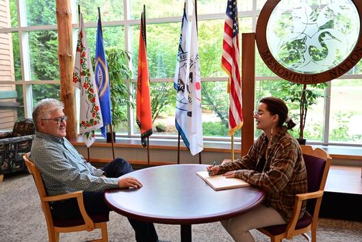 Chris Caldwell, president of College of Menominee Nation, sits with a student on campus. They are at a round table sitting in front of various flags.