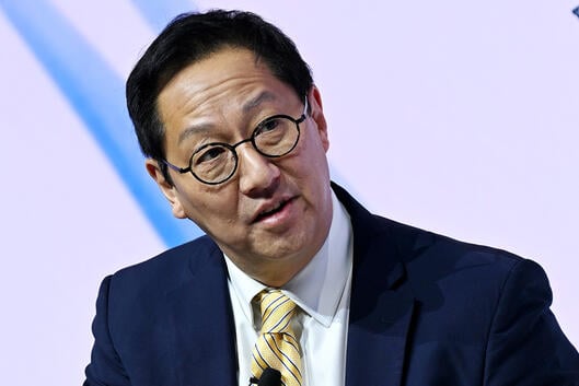 Santa Ono, an Asian man with black hair, wearing glasses and a dark suit with a yellow tie.