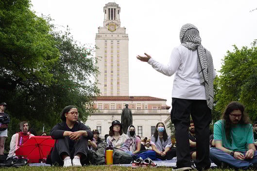 A woman wearing a kaffiyeh as a head scarf addresses a crowd of students seated in the grass in front of a building at the University of Texas at Austin.