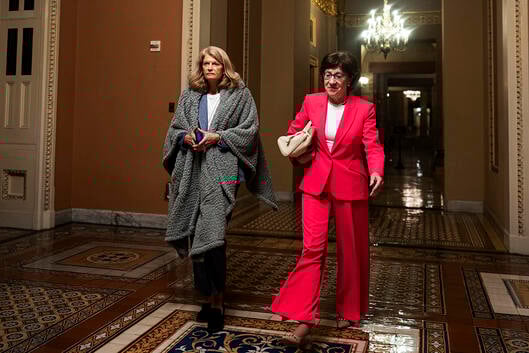 U.S. Sen. Lisa Murkowski (R-AK) and U.S. Sen. Susan Collins (R-ME) walk to the Senate floor during overnight votes at the U.S. Capitol. Murkowski has a blanket wrapped around her shoulders and Collins is wearing a red suit.