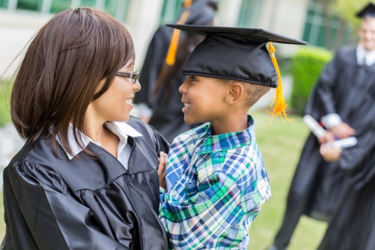 A student in her graduation robe holds her child, who is wearing her mortarboard.