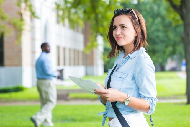 Young woman, presumably on a college tour, smiles as she holds a map in a campus green space.