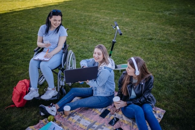 Three female college students sit outside on the grass on a blanket, laughing and looking at a computer screen. One of the women is using a wheelchair.