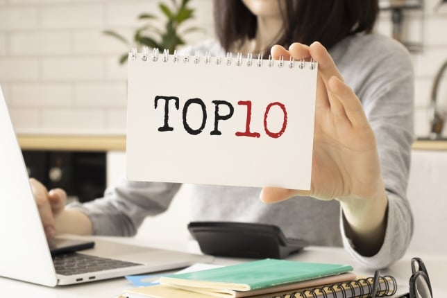 Female student working at a computer at a desk holds up a small spiral notebook that says in bold letters, "Top 10."