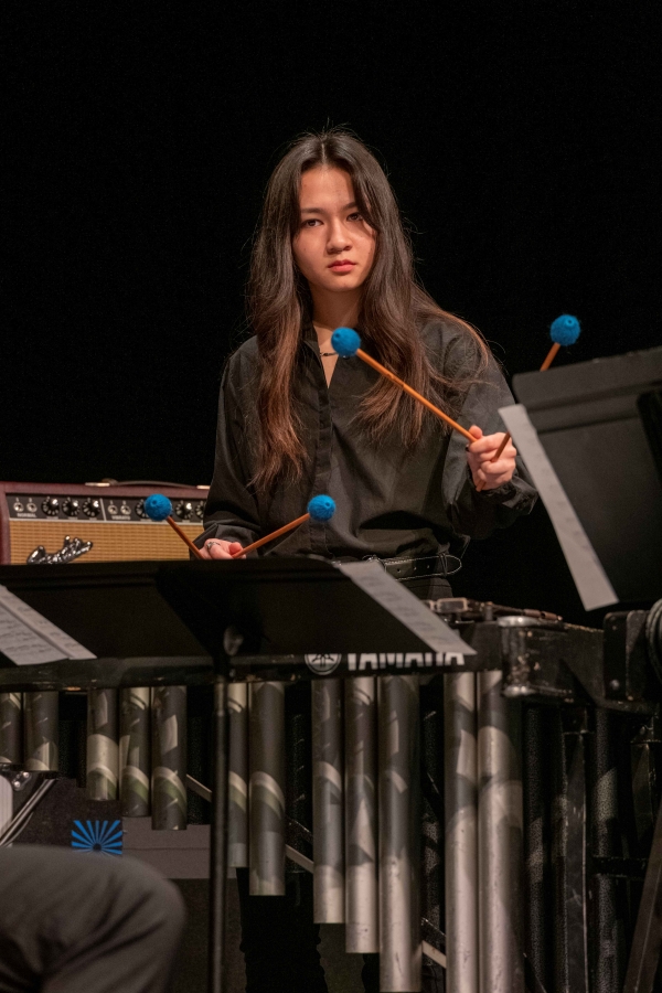 A young woman in a black shirt plays percussion music with mallets on a stage.