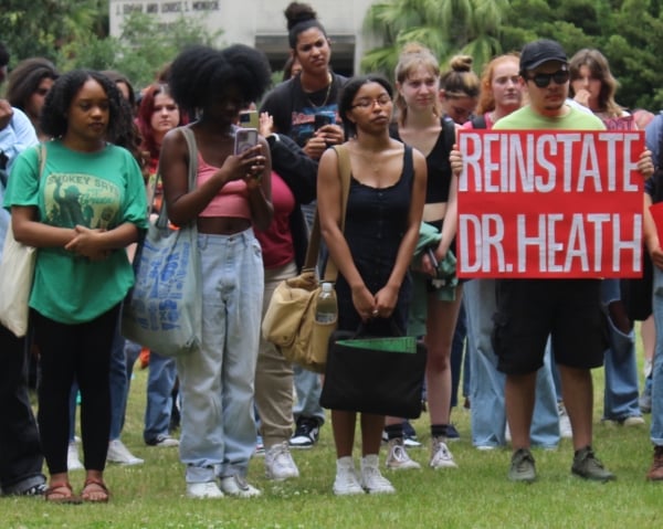 A group of students gather at a demonstration with one of them holding a red sign with white text that reads "Reinstate Dr. Heath"