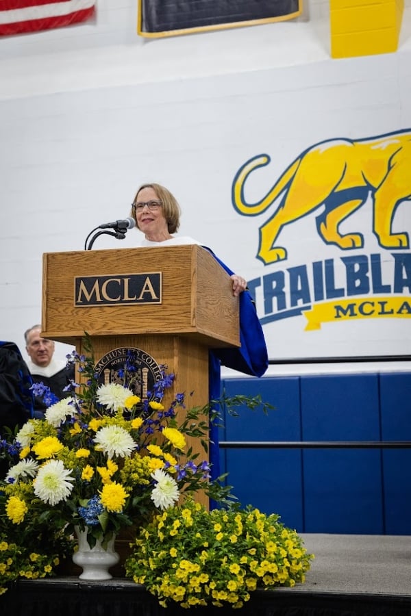 Patricia Okker stands at a podium behind a bouquet of white and yellow flowers and in front of a painting of the MCLA mascot.