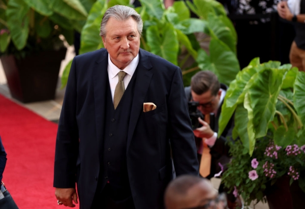A man in a suit and tie on a red carpet surrounded by green plants.