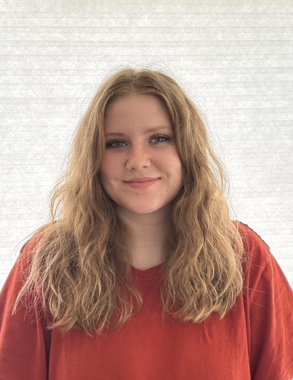 A woman with blonde hair stands in front of a white wall wearing an orange top.