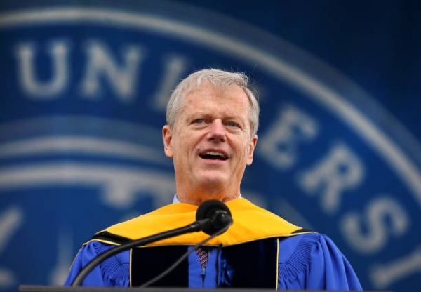 Charlie Baker, wearing a blue robe with a gold hood, speaks at a microphone.
