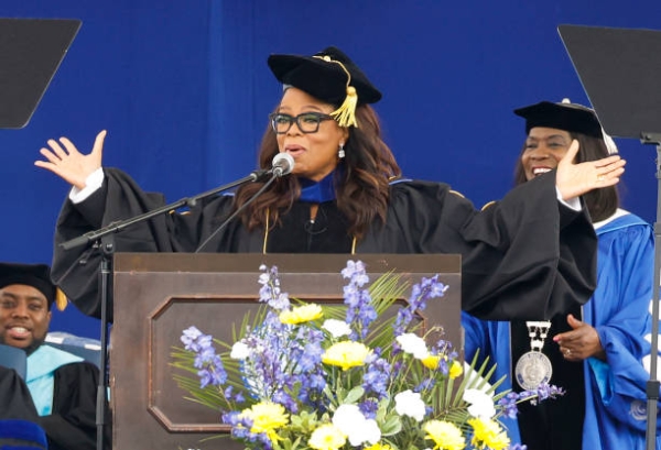 Oprah Winfrey, in a black graduation cap and gown and with her hands outstretched, stands at a podium speaking to the graduates of Tennessee State University.