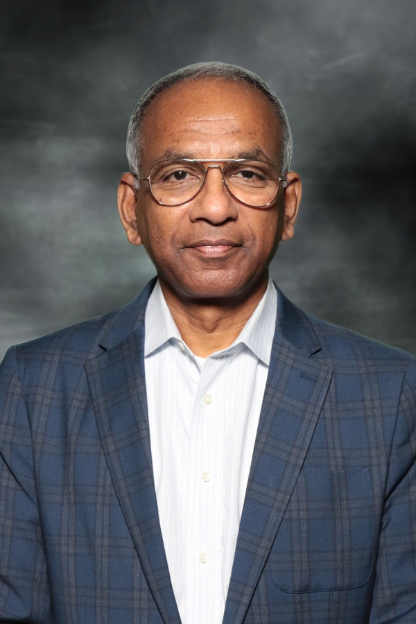 A photo of Johnson Varkey, a brown-skinned man with short gray hair wearing a checked jacket over an open-collared shirt and glasses.