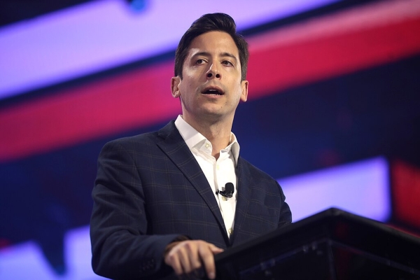 Michael Knowles, a white man with dark hair, speaks in front of a purple and red background. 