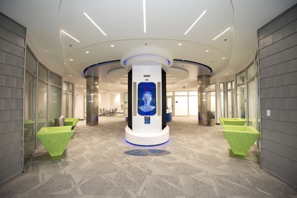 A high-tech holographic concierge—a faceless head and neck, on a blue background—is the centerpiece of a lobby.