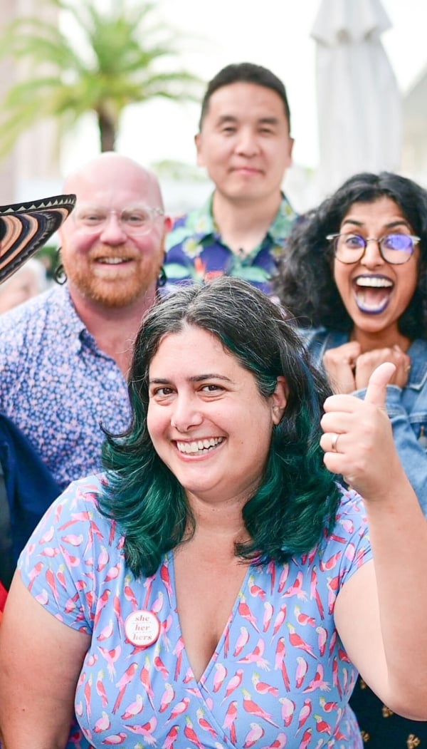 One person in the foreground, a woman with green ombre hair giving a thumbs up, surrounded by three people in the background.