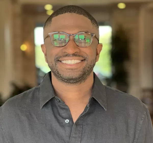 Tade Oyerinde, a young brown-skinned man with short dark hair and a beard wearing glasses and a button-up shirt.