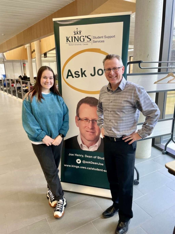 Dean of Students Joe Henry smiles for a photo near his Ask Joe sign with a student