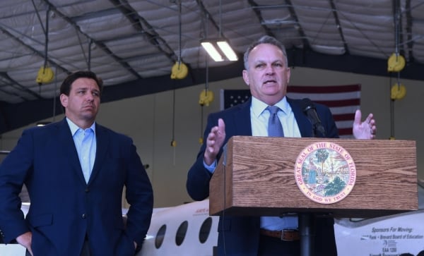 A photo of Ron DeSantis and Richard Corcoran, both wearing dark suits, stand indoors in front of a small airplane. Corcoran is behind a podium.