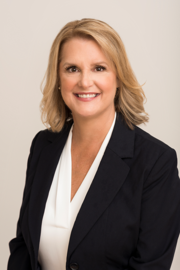 Sherry Hartnett, a light-skinned woman with blond hair, smiles for a headshot wearing a white shirt and a black blazer.