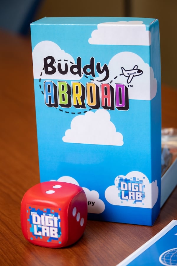 The Buddy Abroad board game box with a DIGI Lab die sit on a table