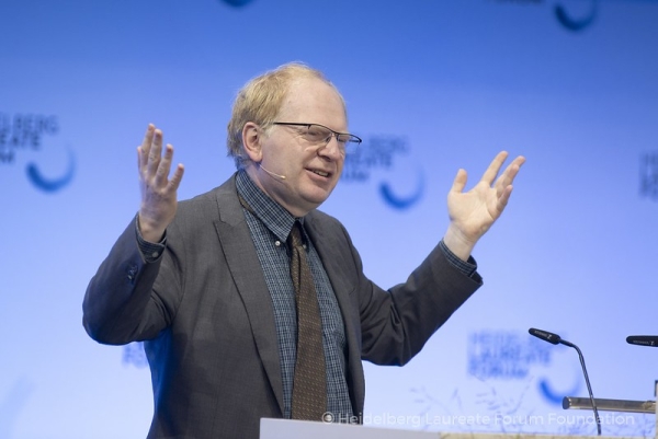 A light-skinned man man with reddish hair in a gray suit and tie stands at a podium giving a lecture with his hands in the air.