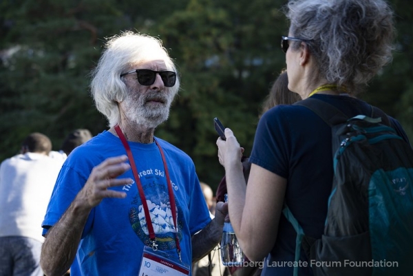 An older man with white hair wearing a blue T-shirt and red lanyard speaks with a reporter whose face the viewer cannot see.