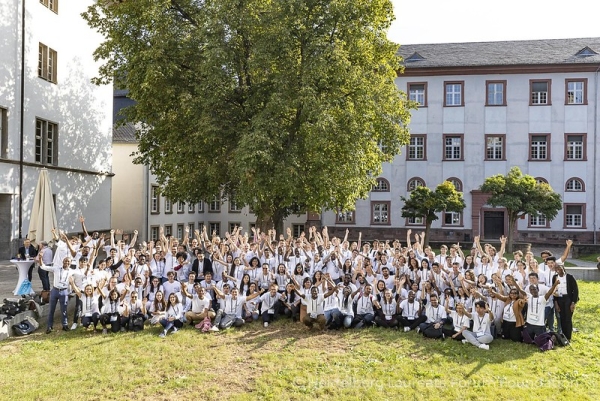 A large, outdoor gathering of young students with raised hands wearing white T-shirts.