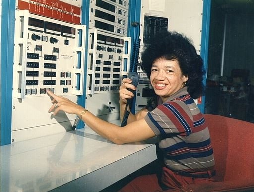 A photo of a smiling woman with light-brown skin and dark hair in front of a dated computer. 