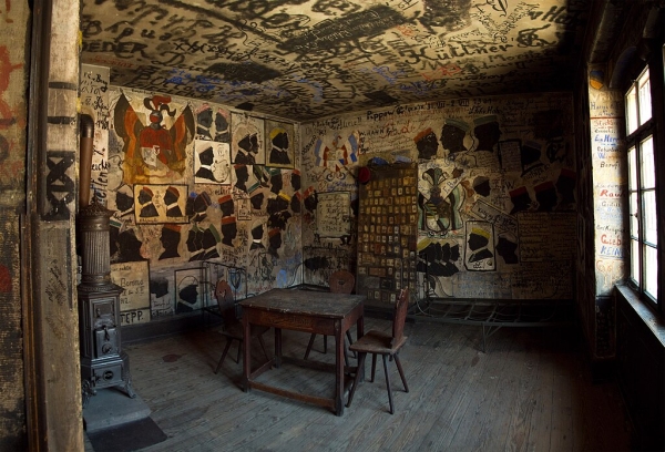 A dim room with a wooden floor, table and chair. The walls are covered in writings and art that look like graffiti.