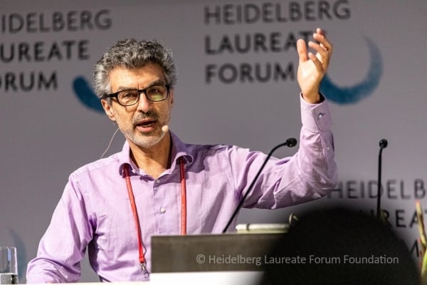 A man with tan skin and graying hair in a pale-purple shirt gives a lecture at a podium with one hand up in the air.