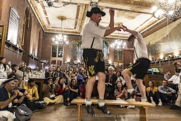 Two people in traditional German attire, including lederhosen and shorts, dance as an audience looks on.