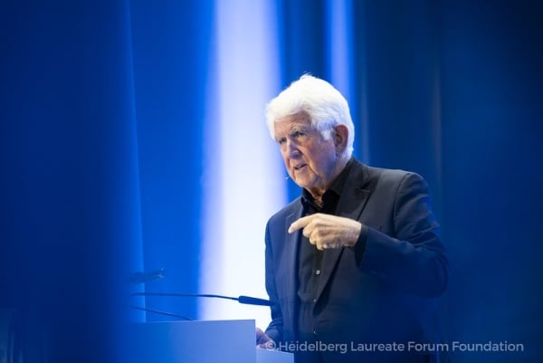 An older white man with white hair stands at a podium and gives a lecture.